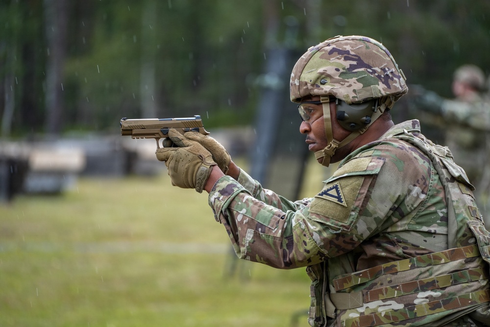 A U.S. Army soldier in full combat gear is aiming a pistol at a firing range during what appears to be a training exercise. The soldier is wearing OCP (Operational Camouflage Pattern) uniform, a combat helmet, hearing protection, eye protection, tactical gloves, and a plate carrier vest. Rain is visibly falling during the exercise.