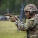 A U.S. Army soldier in full combat gear is aiming a pistol at a firing range during what appears to be a training exercise. The soldier is wearing OCP (Operational Camouflage Pattern) uniform, a combat helmet, hearing protection, eye protection, tactical gloves, and a plate carrier vest. Rain is visibly falling during the exercise.