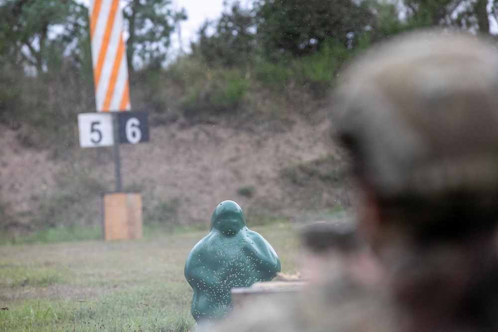 The image shows a military or law enforcement shooting range with a green humanoid target dummy in the foreground showing visible impact marks from projectiles. A person wearing a military-style helmet is partially visible in the right foreground, observing or participating in the exercise. Lane markers numbered 5 and 6 are visible in the background along with an orange and white striped range marker.