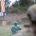 The image shows a military or law enforcement shooting range with a green humanoid target dummy in the foreground showing visible impact marks from projectiles. A person wearing a military-style helmet is partially visible in the right foreground, observing or participating in the exercise. Lane markers numbered 5 and 6 are visible in the background along with an orange and white striped range marker.
