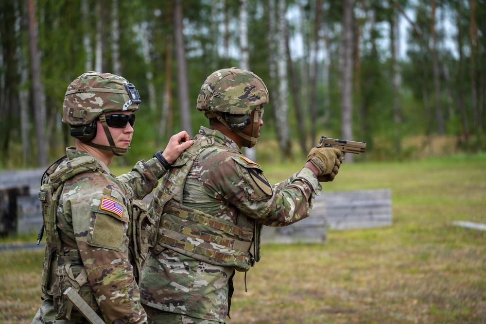 Two U.S. Army soldiers in full combat gear are at an outdoor firing range. One soldier is actively aiming a pistol downrange while the other, wearing sunglasses and hearing protection, appears to be observing or coaching. The setting is a wooded training area with concrete target barriers visible in the background.
