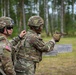 Two U.S. Army soldiers in full combat gear are at an outdoor firing range. One soldier is actively aiming a pistol downrange while the other, wearing sunglasses and hearing protection, appears to be observing or coaching. The setting is a wooded training area with concrete target barriers visible in the background.