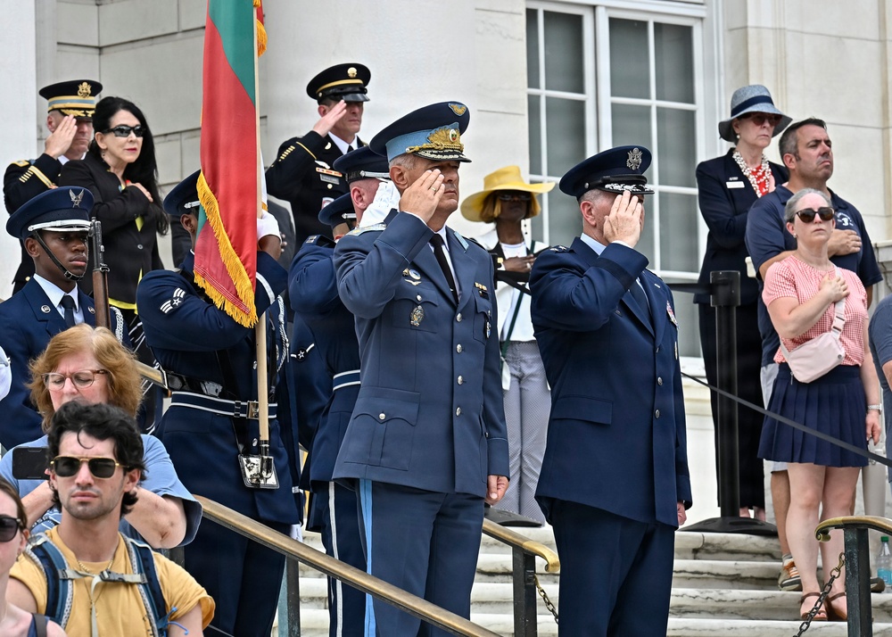 A formal military ceremony is taking place on the steps of what appears to be a government or memorial building. Two senior officers in blue air force dress uniforms are rendering salutes, accompanied by honor guard members and civilians observing the ceremony. A flag with red and green coloring (consistent with the Lithuanian or Portuguese flag) is visible being carried by a color guard member.