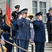 A formal military ceremony is taking place on the steps of what appears to be a government or memorial building. Two senior officers in blue air force dress uniforms are rendering salutes, accompanied by honor guard members and civilians observing the ceremony. A flag with red and green coloring (consistent with the Lithuanian or Portuguese flag) is visible being carried by a color guard member.