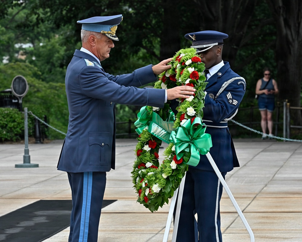Two military personnel are participating in a formal wreath-laying ceremony at what appears to be a memorial site, likely Arlington National Cemetery. A senior officer in a light blue foreign military uniform and a U.S. Air Force honor guard member in dress blues are jointly handling a large decorative wreath with red, white, and green flowers and a green ribbon. A civilian observer is visible in the background.