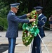 Two military personnel are participating in a formal wreath-laying ceremony at what appears to be a memorial site, likely Arlington National Cemetery. A senior officer in a light blue foreign military uniform and a U.S. Air Force honor guard member in dress blues are jointly handling a large decorative wreath with red, white, and green flowers and a green ribbon. A civilian observer is visible in the background.