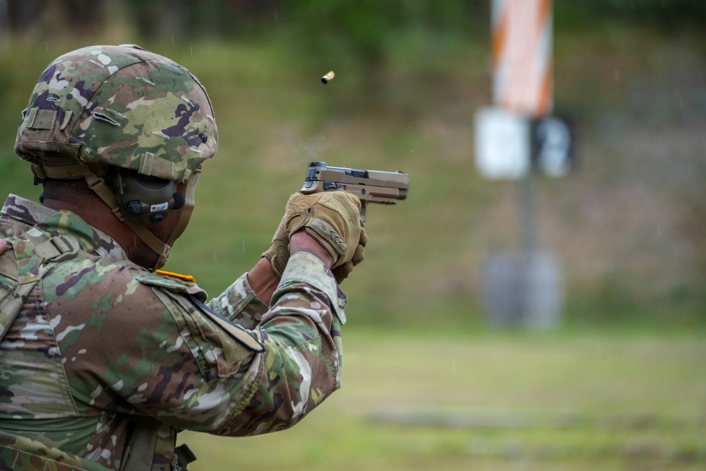 A soldier in full combat gear is firing a pistol at a shooting range, with a spent brass casing visibly ejecting from the firearm. The individual is wearing MultiCam/OCP camouflage uniform, a combat helmet, face covering, and tactical gloves. A silhouette target is visible in the blurred background.