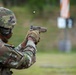 A soldier in full combat gear is firing a pistol at a shooting range, with a spent brass casing visibly ejecting from the firearm. The individual is wearing MultiCam/OCP camouflage uniform, a combat helmet, face covering, and tactical gloves. A silhouette target is visible in the blurred background.