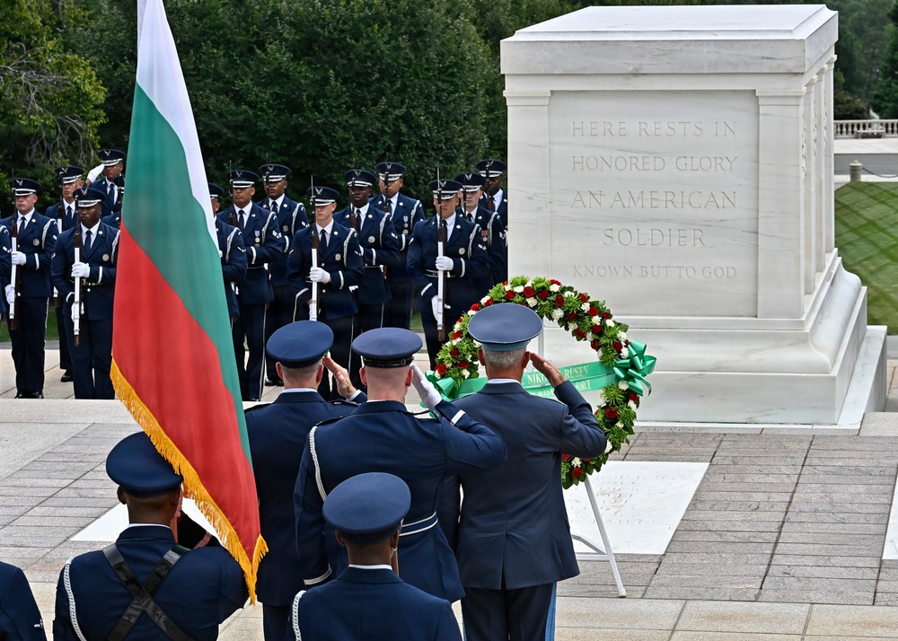 A formal military wreath-laying ceremony is taking place at the Tomb of the Unknown Soldier at Arlington National Cemetery. Military personnel in dress uniforms are saluting while a Bulgarian flag is prominently displayed, suggesting an official visit by Bulgarian military or government officials. A floral wreath with red, white, and green flowers and a green ribbon is placed on a stand before the marble tomb.
