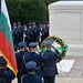 A formal military wreath-laying ceremony is taking place at the Tomb of the Unknown Soldier at Arlington National Cemetery. Military personnel in dress uniforms are saluting while a Bulgarian flag is prominently displayed, suggesting an official visit by Bulgarian military or government officials. A floral wreath with red, white, and green flowers and a green ribbon is placed on a stand before the marble tomb.