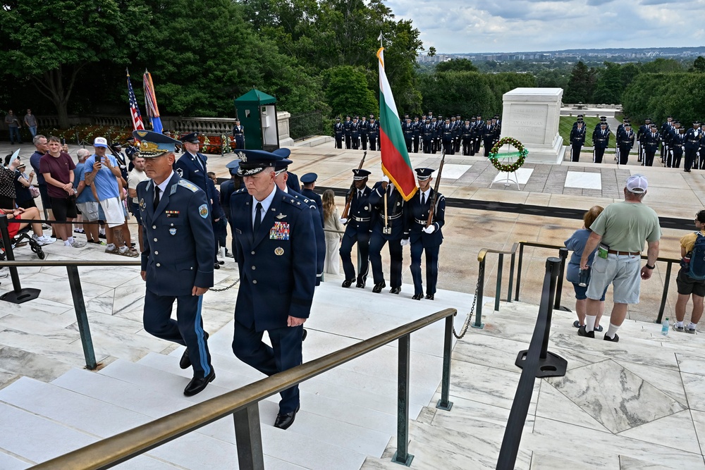 A formal military wreath-laying ceremony is taking place at the Tomb of the Unknown Soldier at Arlington National Cemetery, Virginia. Two senior military officers in dress uniforms — one appearing to be a foreign dignitary carrying a Bulgarian flag — are descending the marble steps following the ceremony. A formation of U.S. Air Force honor guard personnel stands at attention near a wreath placed at the tomb.