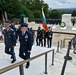 A formal military wreath-laying ceremony is taking place at the Tomb of the Unknown Soldier at Arlington National Cemetery, Virginia. Two senior military officers in dress uniforms — one appearing to be a foreign dignitary carrying a Bulgarian flag — are descending the marble steps following the ceremony. A formation of U.S. Air Force honor guard personnel stands at attention near a wreath placed at the tomb.