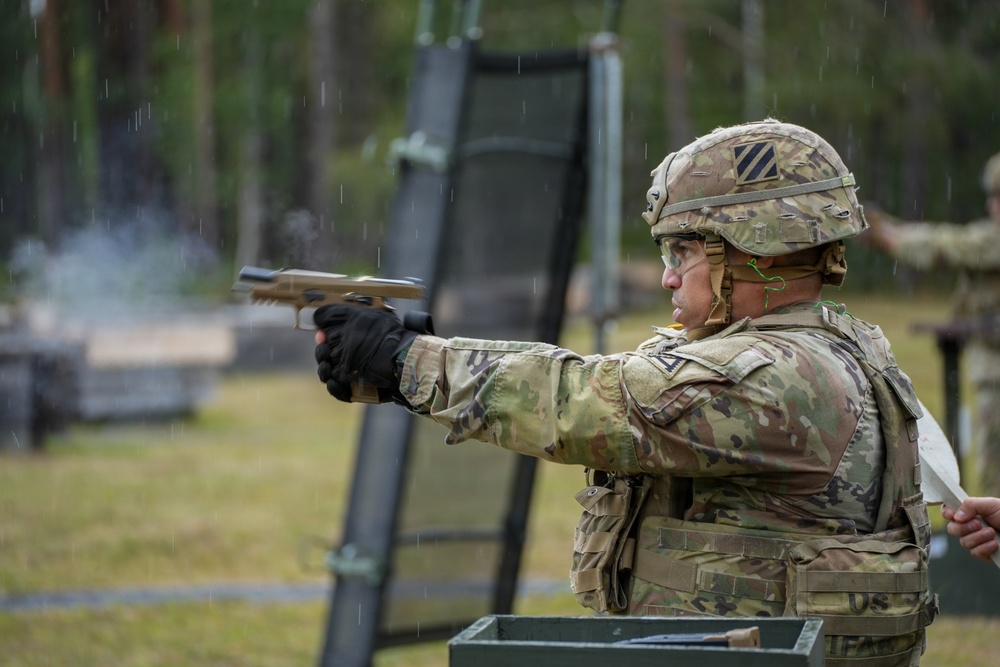 A U.S. Army soldier in full combat gear is firing a pistol at an outdoor shooting range during rainy conditions. The soldier wears a 3rd Infantry Division patch on their helmet and Operational Camouflage Pattern (OCP) uniform with body armor. The image captures an active weapons qualification or training exercise.