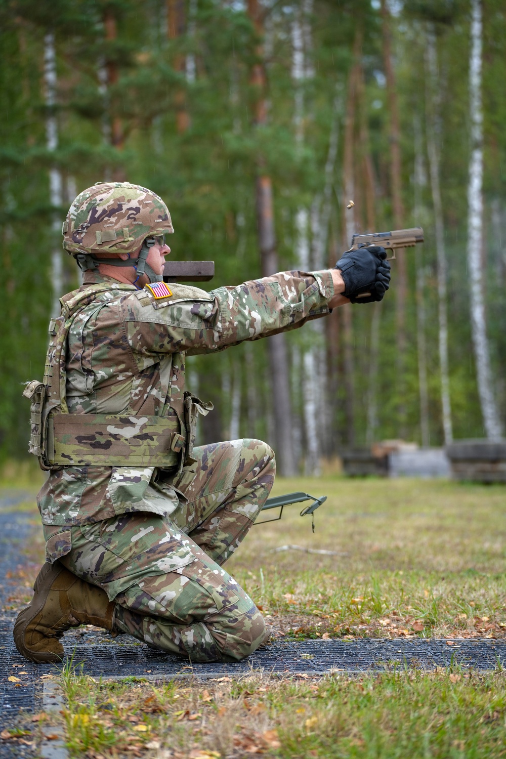 A U.S. Army soldier in full combat gear is firing a pistol from a kneeling position on what appears to be a military training range. The soldier is wearing Operational Camouflage Pattern (OCP) uniform with a visible American flag patch on the left shoulder, a combat helmet, body armor, and tactical gloves. A spent brass casing is visibly ejecting from the pistol, indicating the weapon has just been fired.