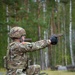 A U.S. Army soldier in full combat gear is firing a pistol from a kneeling position on what appears to be a military training range. The soldier is wearing Operational Camouflage Pattern (OCP) uniform with a visible American flag patch on the left shoulder, a combat helmet, body armor, and tactical gloves. A spent brass casing is visibly ejecting from the pistol, indicating the weapon has just been fired.