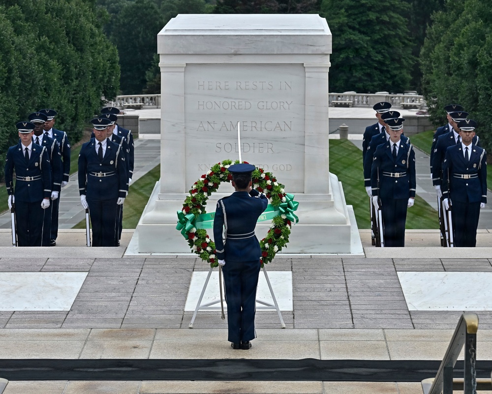 A formal military wreath-laying ceremony is taking place at the Tomb of the Unknown Soldier at Arlington National Cemetery. A single service member stands at attention before a floral wreath on a stand, facing the marble tomb inscribed 'HERE RESTS IN HONORED GLORY AN AMERICAN SOLDIER KNOWN BUT TO GOD.' Multiple uniformed military personnel, appearing to be U.S. Air Force honor guard members, are positioned symmetrically on both sides of the tomb.