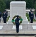 A formal military wreath-laying ceremony is taking place at the Tomb of the Unknown Soldier at Arlington National Cemetery. A single service member stands at attention before a floral wreath on a stand, facing the marble tomb inscribed 'HERE RESTS IN HONORED GLORY AN AMERICAN SOLDIER KNOWN BUT TO GOD.' Multiple uniformed military personnel, appearing to be U.S. Air Force honor guard members, are positioned symmetrically on both sides of the tomb.