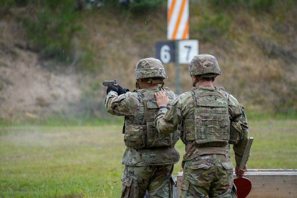 Two military personnel in full combat gear are at an outdoor shooting range during rainy conditions. One soldier is actively firing a pistol downrange while the other, likely an instructor or range safety officer, stands alongside observing. Lane markers numbered 6 and 7 are visible in the background.