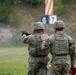 Two military personnel in full combat gear are at an outdoor shooting range during rainy conditions. One soldier is actively firing a pistol downrange while the other, likely an instructor or range safety officer, stands alongside observing. Lane markers numbered 6 and 7 are visible in the background.