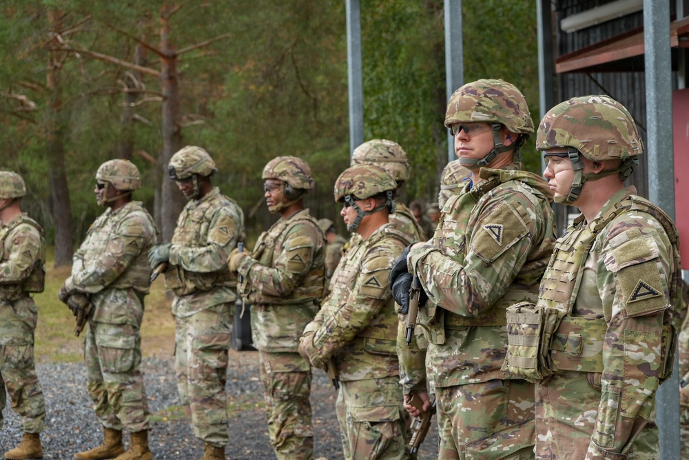 A group of U.S. Army soldiers in full combat gear are standing in a formation line outdoors, likely at a training facility or military base. They are wearing OCP (Operational Camouflage Pattern) uniforms, tactical helmets, and body armor. The setting appears to be a wooded military training area with gravel ground and a structure visible on the right.