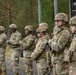 A group of U.S. Army soldiers in full combat gear are standing in a formation line outdoors, likely at a training facility or military base. They are wearing OCP (Operational Camouflage Pattern) uniforms, tactical helmets, and body armor. The setting appears to be a wooded military training area with gravel ground and a structure visible on the right.