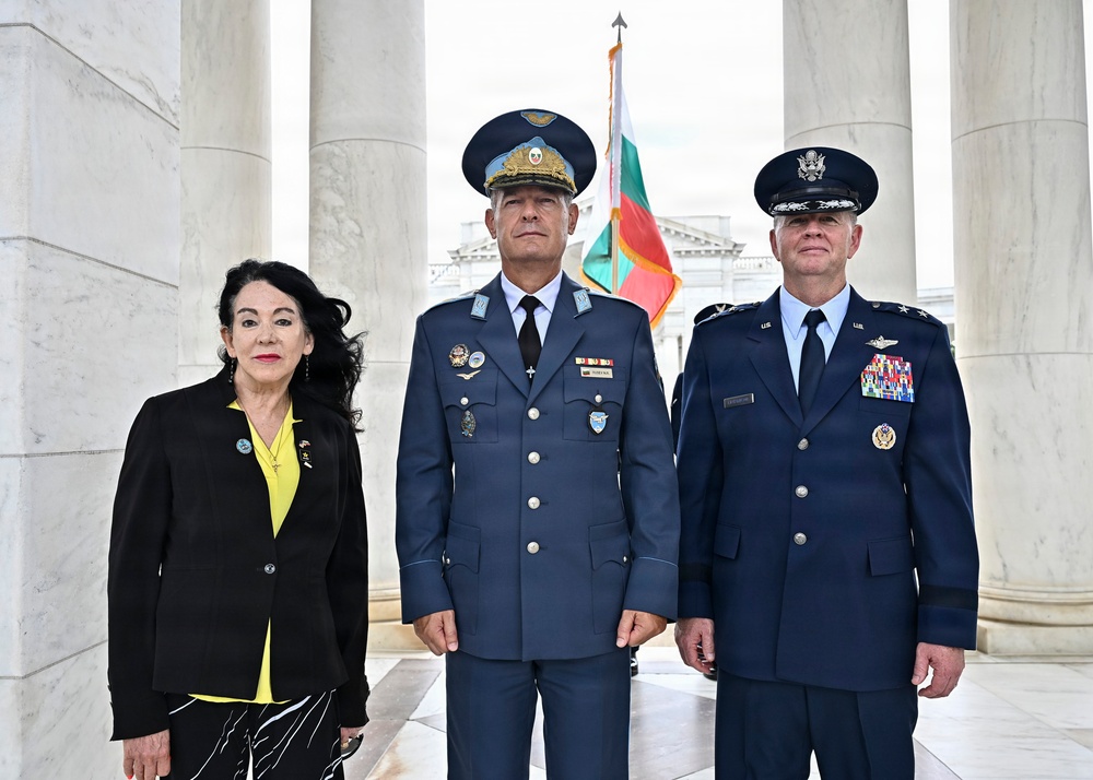 Three individuals pose formally between marble columns, likely at a memorial or government building. Two military officers in dress uniforms flank a civilian woman in a black jacket with yellow accents. A flag consistent with the Bulgarian tricolor is visible in the background.