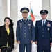 Three individuals pose formally between marble columns, likely at a memorial or government building. Two military officers in dress uniforms flank a civilian woman in a black jacket with yellow accents. A flag consistent with the Bulgarian tricolor is visible in the background.