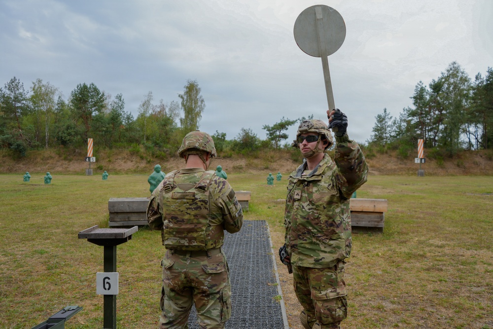 Two soldiers in US Army MultiCam (OCP) camouflage uniforms are at a military shooting range, standing at lane number 6. One soldier is holding a circular paddle/disc raised in the air, likely signaling a range command, while the other faces downrange. Multiple green humanoid silhouette targets and numbered lane markers are visible in the background.