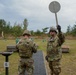 Two soldiers in US Army MultiCam (OCP) camouflage uniforms are at a military shooting range, standing at lane number 6. One soldier is holding a circular paddle/disc raised in the air, likely signaling a range command, while the other faces downrange. Multiple green humanoid silhouette targets and numbered lane markers are visible in the background.
