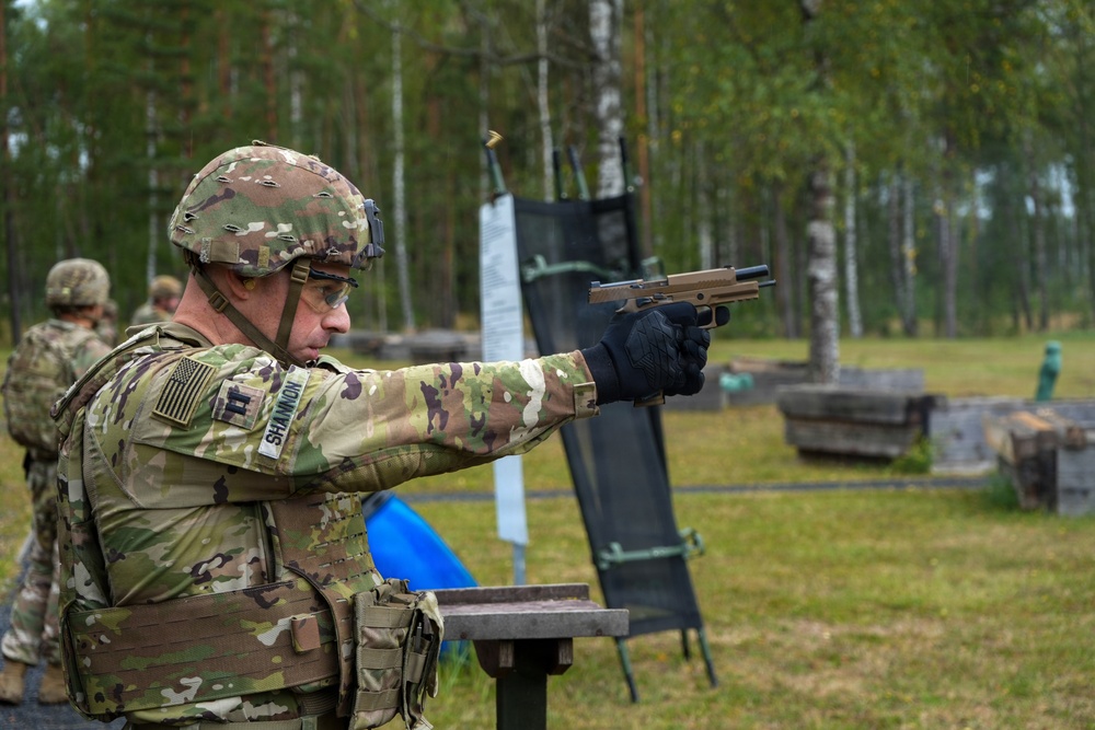 A U.S. Army soldier identified by name tape as 'Shannon' is firing a pistol at a military shooting range. The soldier is wearing full combat gear including OCP multicam uniform, tactical helmet, body armor, and black gloves. A spent brass casing is visible being ejected from the firearm, indicating an active firing moment.