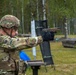 A U.S. Army soldier identified by name tape as 'Shannon' is firing a pistol at a military shooting range. The soldier is wearing full combat gear including OCP multicam uniform, tactical helmet, body armor, and black gloves. A spent brass casing is visible being ejected from the firearm, indicating an active firing moment.