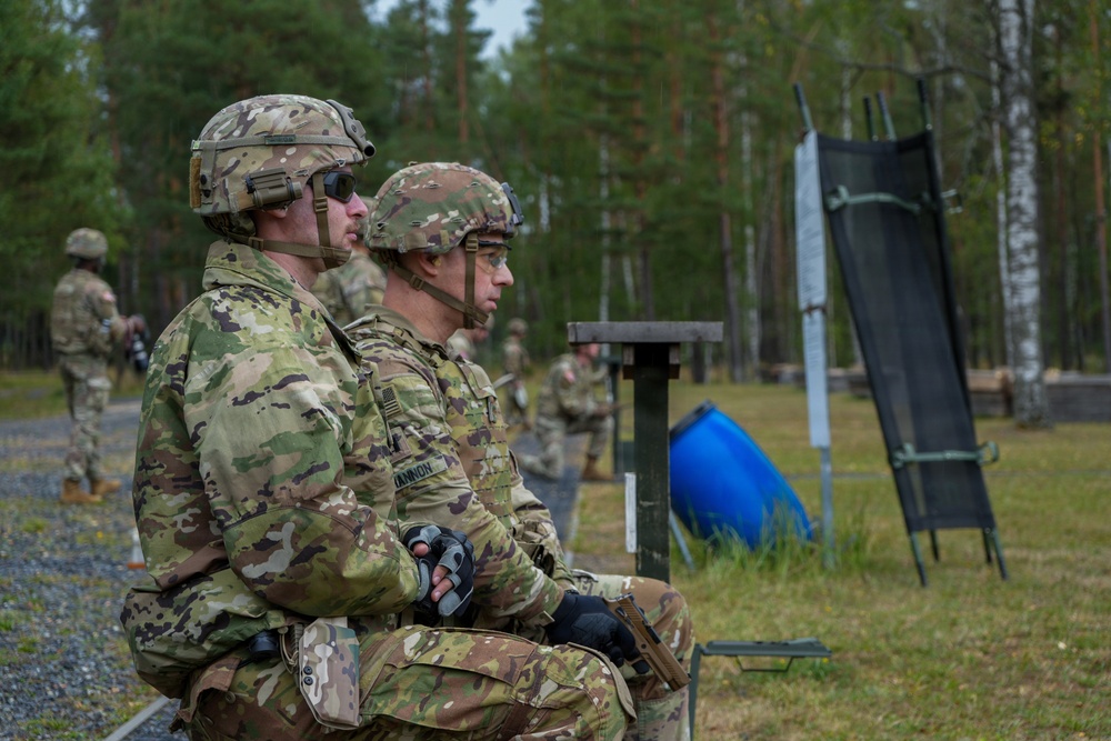 Two U.S. Army soldiers in MultiCam (OCP) uniforms and helmets are seated at what appears to be a military firing range, observing downrange activity. A shooting target stand with a dark panel is visible to their right, and additional soldiers can be seen in the background. The setting appears to be a forested training area, likely in Europe based on the pine tree landscape.