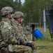 Two U.S. Army soldiers in MultiCam (OCP) uniforms and helmets are seated at what appears to be a military firing range, observing downrange activity. A shooting target stand with a dark panel is visible to their right, and additional soldiers can be seen in the background. The setting appears to be a forested training area, likely in Europe based on the pine tree landscape.