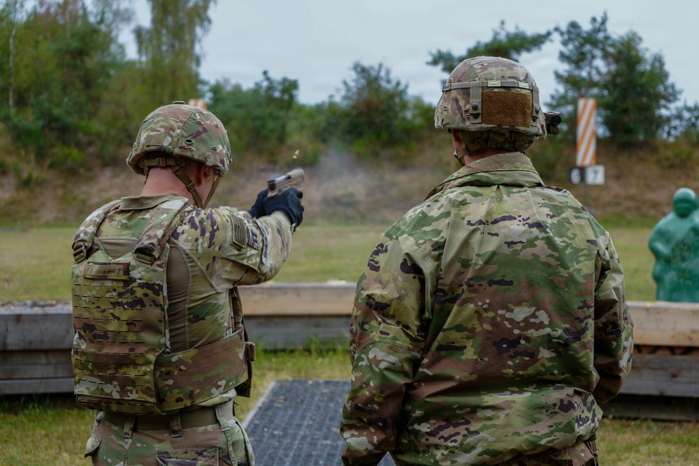 Two military personnel in full combat gear are at an outdoor firing range. The soldier on the left is actively firing a pistol downrange, with visible muzzle smoke, while the soldier on the right observes. The setting appears to be a standard military qualification or training range with target silhouettes visible in the background.