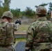 Two military personnel in full combat gear are at an outdoor firing range. The soldier on the left is actively firing a pistol downrange, with visible muzzle smoke, while the soldier on the right observes. The setting appears to be a standard military qualification or training range with target silhouettes visible in the background.