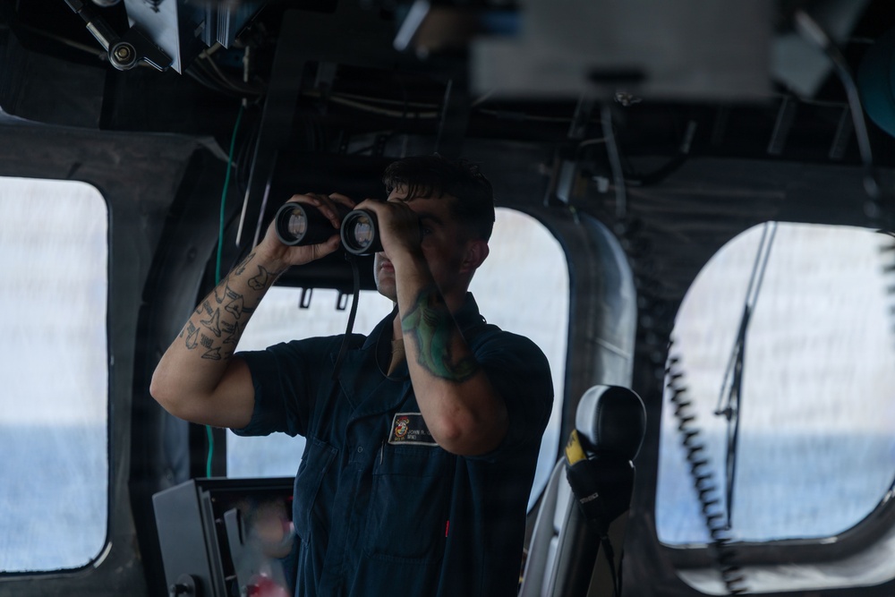 A naval service member in dark blue coveralls stands on what appears to be a ship's bridge, looking through binoculars. The individual has visible tattoos on both forearms and wears a name tag and unit patch on their uniform. The setting is clearly a military vessel's pilothouse or bridge wing, with ocean visible through the windows.