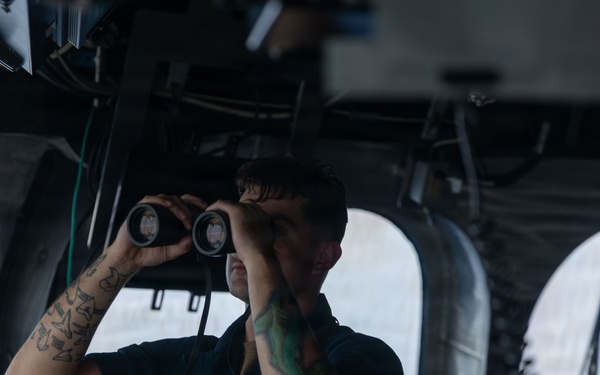 A naval service member in dark blue coveralls stands on what appears to be a ship's bridge, looking through binoculars. The individual has visible tattoos on both forearms and wears a name tag and unit patch on their uniform. The setting is clearly a military vessel's pilothouse or bridge wing, with ocean visible through the windows.