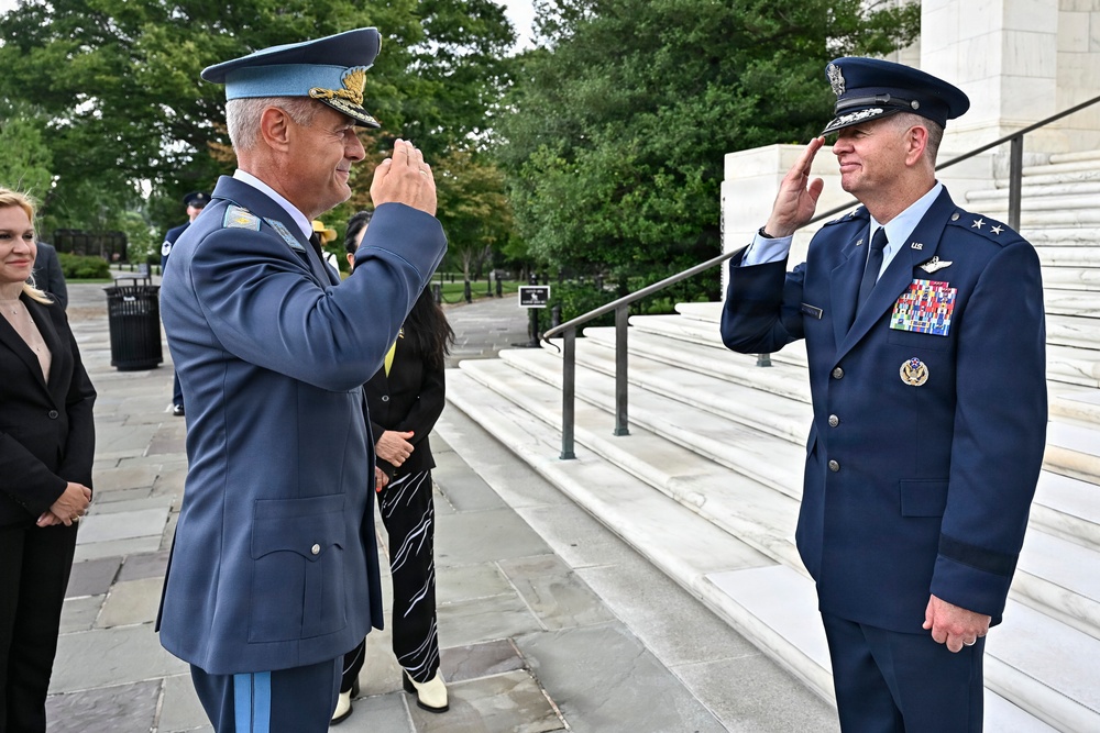 Two senior military officers in dress uniforms are exchanging salutes in what appears to be a formal ceremonial setting, likely at Arlington National Cemetery based on the white marble architecture visible in the background. One officer wears a lighter blue uniform with a distinctive peaked cap suggesting a foreign air force, while the other wears a US Air Force dress uniform with two-star general insignia. Several civilians are observing the exchange in the background.