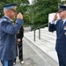Two senior military officers in dress uniforms are exchanging salutes in what appears to be a formal ceremonial setting, likely at Arlington National Cemetery based on the white marble architecture visible in the background. One officer wears a lighter blue uniform with a distinctive peaked cap suggesting a foreign air force, while the other wears a US Air Force dress uniform with two-star general insignia. Several civilians are observing the exchange in the background.