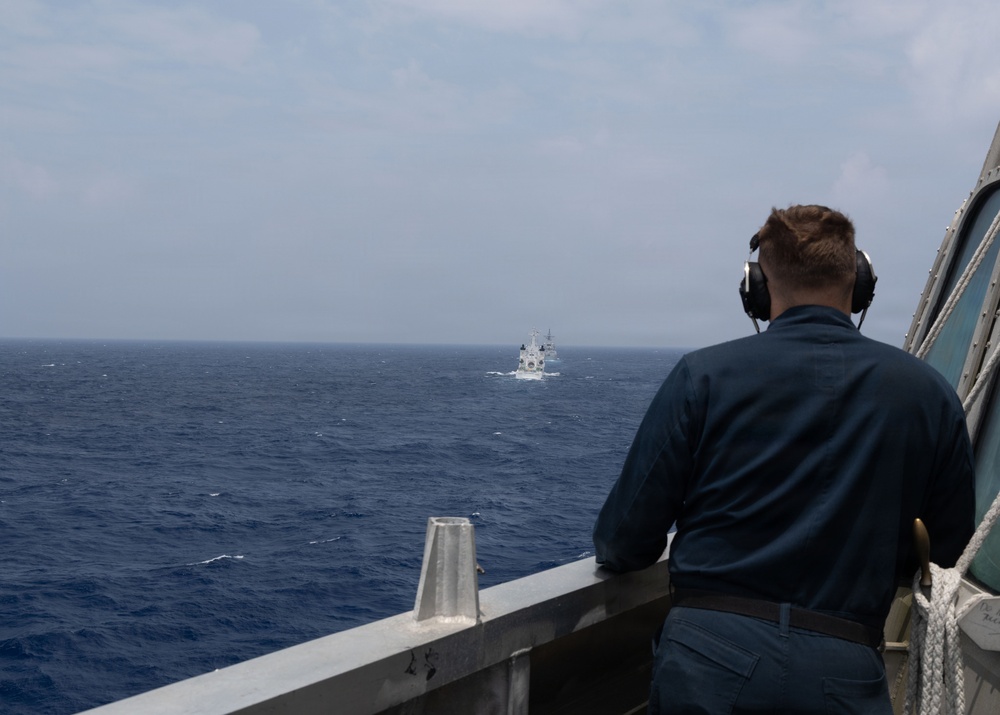 A naval personnel member wearing communication headphones stands on the deck of a warship, observing another vessel in the distance across open ocean. The observer is positioned at a railing on what appears to be a bridge wing or observation deck. The distant vessel appears to be a white-hulled ship, possibly a naval or coast guard vessel.