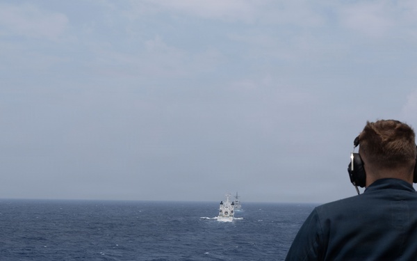 A naval personnel member wearing communication headphones stands on the deck of a warship, observing another vessel in the distance across open ocean. The observer is positioned at a railing on what appears to be a bridge wing or observation deck. The distant vessel appears to be a white-hulled ship, possibly a naval or coast guard vessel.