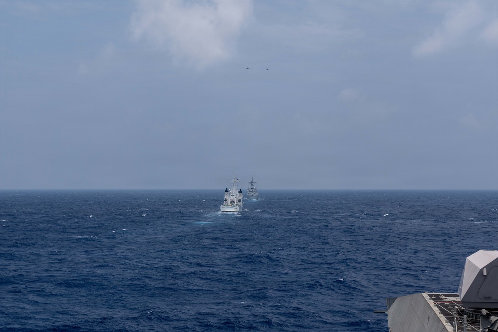 A naval formation is underway at sea, showing multiple warships sailing in column formation across open ocean. The image is taken from the deck of a warship in the foreground, looking toward two additional vessels in the middle distance. At least one or two aircraft are visible flying at high altitude above the formation.