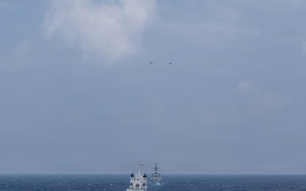 A naval formation is underway at sea, showing multiple warships sailing in column formation across open ocean. The image is taken from the deck of a warship in the foreground, looking toward two additional vessels in the middle distance. At least one or two aircraft are visible flying at high altitude above the formation.