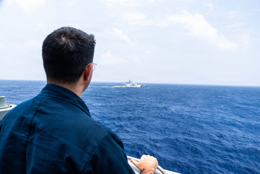 A person wearing a dark navy uniform and glasses is observed from behind, standing on the deck of a vessel and looking out toward another ship in the distance on open blue ocean waters. The distant vessel appears to be a large white coast guard or patrol ship. The scene suggests a maritime observation or monitoring situation.