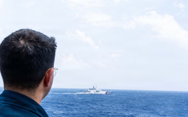 A person wearing a dark navy uniform and glasses is observed from behind, standing on the deck of a vessel and looking out toward another ship in the distance on open blue ocean waters. The distant vessel appears to be a large white coast guard or patrol ship. The scene suggests a maritime observation or monitoring situation.