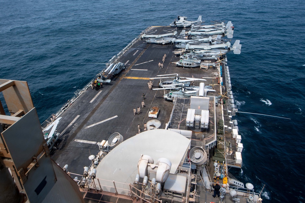 An aerial view of a large amphibious assault ship's flight deck taken from an elevated position, likely the island superstructure. The deck is populated with multiple military aircraft including tiltrotor V-22 Ospreys and helicopters, along with deck crew personnel conducting operations. The vessel is underway at sea with calm, deep blue ocean visible on both sides.