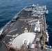 An aerial view of a large amphibious assault ship's flight deck taken from an elevated position, likely the island superstructure. The deck is populated with multiple military aircraft including tiltrotor V-22 Ospreys and helicopters, along with deck crew personnel conducting operations. The vessel is underway at sea with calm, deep blue ocean visible on both sides.