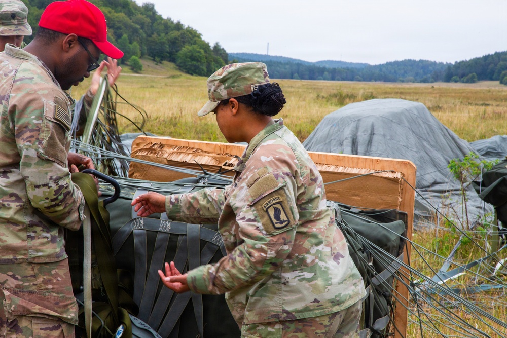 Two U.S. Army soldiers in OCP (Operational Camouflage Pattern) uniforms are working with airdrop equipment in an open field. The female soldier displays an Airborne tab and 173rd Airborne Brigade patch on her left shoulder. They appear to be recovering or inspecting a cargo airdrop bundle with parachute suspension lines after a drop.