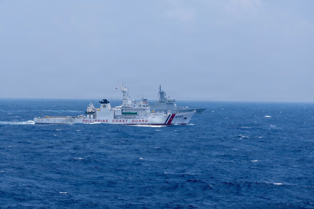 The image shows a Philippine Coast Guard vessel (hull number 9701) sailing in open, choppy blue waters under an overcast sky. A second, larger gray warship is visible in the background, partially obscured by haze. The scene appears to depict two vessels operating in close proximity at sea, likely in the South China Sea region.