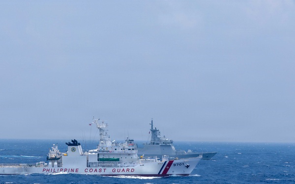 The image shows a Philippine Coast Guard vessel (hull number 9701) sailing in open, choppy blue waters under an overcast sky. A second, larger gray warship is visible in the background, partially obscured by haze. The scene appears to depict two vessels operating in close proximity at sea, likely in the South China Sea region.